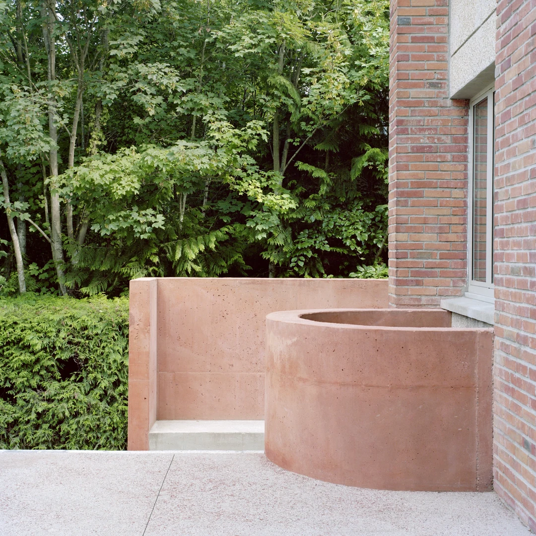 Bassin circulaire en béton rose adossé à une façade de brique, devant un massif boisé