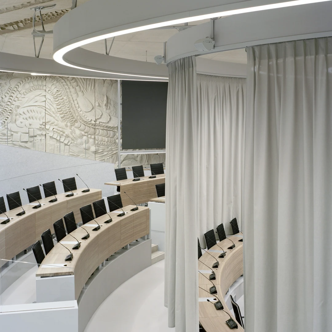 Hémicycle aux pupitres en bois clair et rideaux blancs sous un anneau lumineux